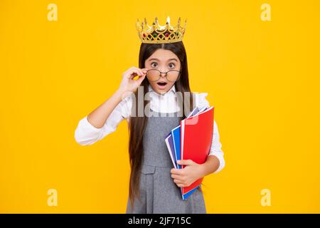 Surprised teenager girl. Schoolgirl in school uniform and crown celebrating victory on yellow background. School child hold books. Education Stock Photo