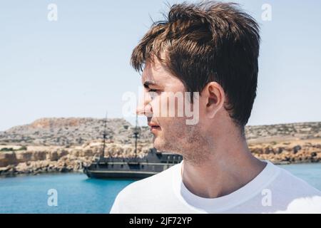 Portrait of a man against the background of a tourist ship, a yellow stony beach and a blue sea. Cape Greco Peninsula Park, Cyprus. The guy looks away. Travel rest. Copy space. Stock Photo