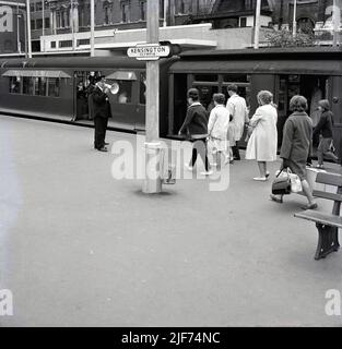 1960s, historical, rail passengers walking to board the District line at Kensington Olympia station, Kensington, London, England, UK. A uniformed train guard with megaphone on the platform announcing the departure. A combined rail and tube station, Kensington (Olympia) has been served by a short branch section of the District line since 1946, originally by as part of the Middle Circle, a GWR service in London that operated from 1872 to 1905. The station's name comes from it's location in Kensington and the adjacent Olympia exhibition centre and was originally opened in 1844. Stock Photo