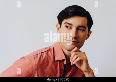 Headshot thoughtful handsome young adult latino man with hand on chin black hair and pink shirt over gray background looking away at copy space studio shot Stock Photo