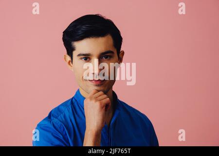 Headshot thoughtful handsome young adult latino man with hand on chin black hair and blue shirt over pink background looking at camera studio shot Stock Photo