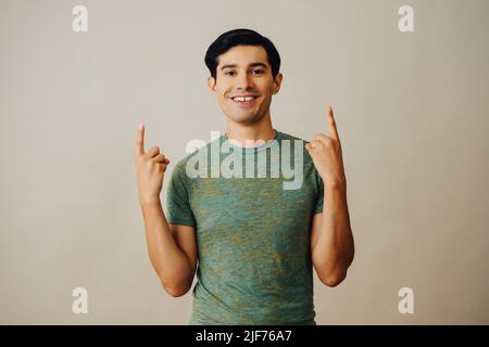 Portrait hispanic latino man pointing up black hair smiling handsome young adult green t-shirt over gray background looking at camera studio shot Stock Photo