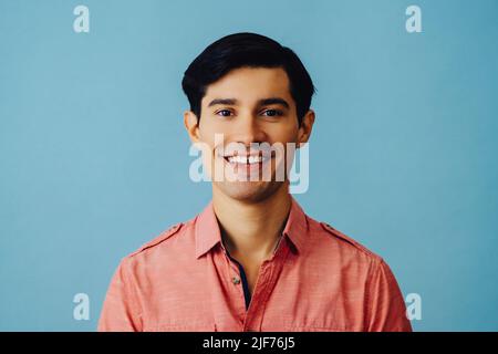 Headshot hispanic latino man black hair smiling handsome young adult wearing pink shirt over blue background looking at camera studio shot Stock Photo