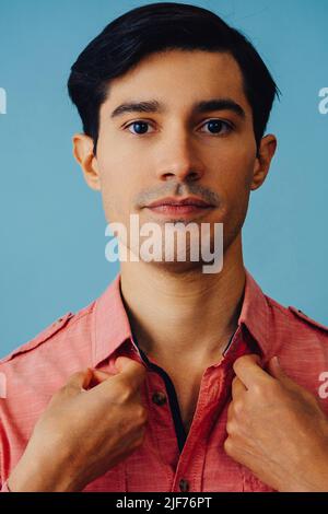 Headshot hispanic latino man black hair smiling handsome young adult wearing pink shirt over blue background looking at camera studio shot Stock Photo