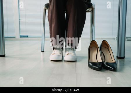 Businesswoman changing uncomfortable black high heels on comfortable white sneakers in office. Stock Photo