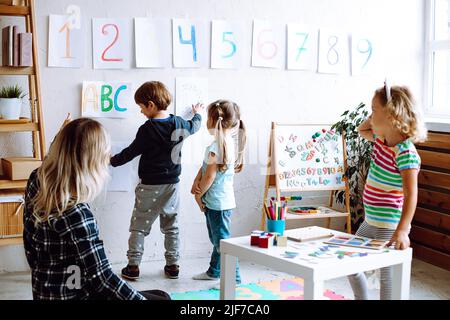 Little girls and educator looking at boy showing pictures on wall in playroom. Interesting logic and intelligence lesson for kindergartners Stock Photo