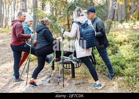 Group of campers stretching, warming up before Nordic walking with trekking sticks in forest, standing one leg on bench Stock Photo