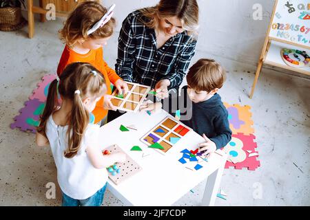 Little kids with educator folding colorful details of constructor on desk in playroom from above view. Interesting game for kindergartners developing Stock Photo