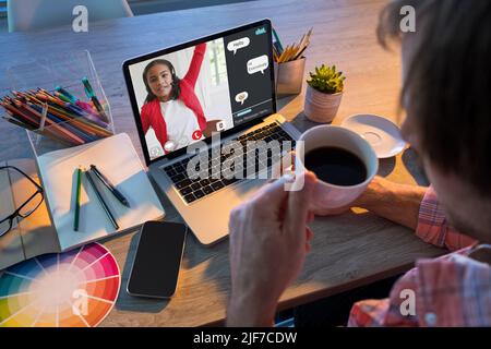 Caucasian male teacher drinking coffee and looking at biracial student raising hand in online class Stock Photo