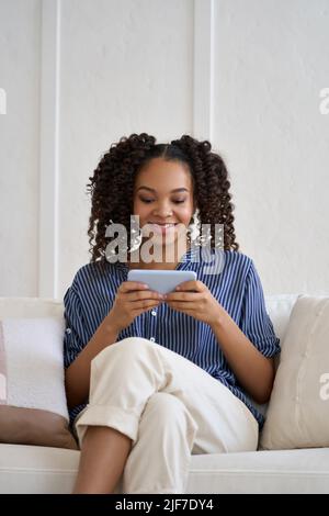 Happy teenage African American girl playing mobile game on phone at home. Stock Photo