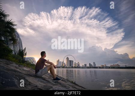 Young man sitting on waterfront and looking at dramatic clouds of thunderstorm above urban skyline, Singapore. Stock Photo