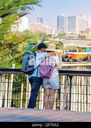 Back view of Asian couple relaxing and chilling at a riverside. Relaxed lifestyle for lover enjoying freedom in summer holidays. Happy people outdoors Stock Photo