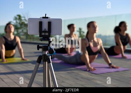 Mobile phone on tripod shooting or streaming group yoga outdoor class online. Stock Photo