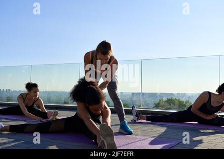 Fit sporty yoga teacher helping african woman on stretching group outdoor class. Stock Photo