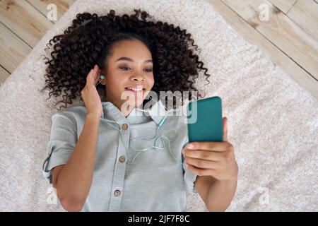 Happy African teen girl in earphones lying in bed listening music on smartphone. Stock Photo