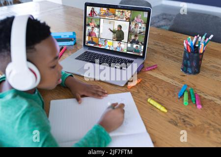 African american boy wearing headphones and listening to online class while writing in book at home Stock Photo