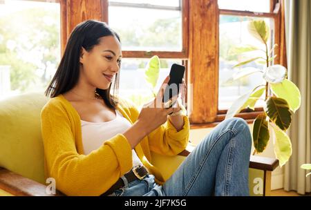 Hispanic woman using her smartphone sitting in front of a window in a bright living room. A young happy female smiling at her cellphone relaxing on a Stock Photo