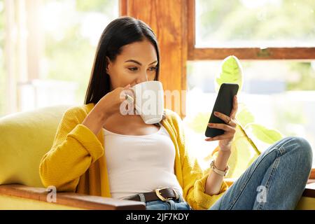 Woman using her smartphone and drinking coffee sitting in front of a window in a bright living room. A young female looking at her cellphone relaxing Stock Photo