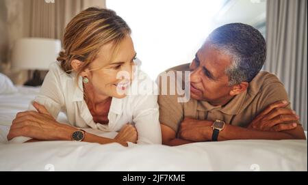 Interracial senior couple relaxing on bed in their modern apartment. Happy mature man and woman on a romantic vacation bonding and talking together in Stock Photo