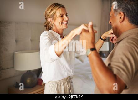 Older interracial couple dancing in a hotel bedroom together on a romantic holiday. Senior hispanic man and caucasian woman smiling and playing at Stock Photo