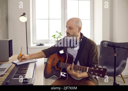 Focused male musician writes in notebook while sitting with guitar in music studio. Stock Photo