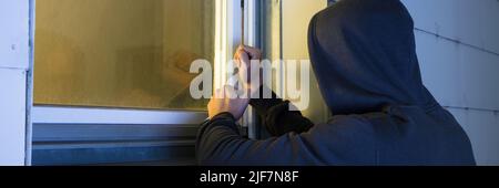 Close-up Of Robber In Hood Using The Red Crowbar To Open The Glass Window Stock Photo