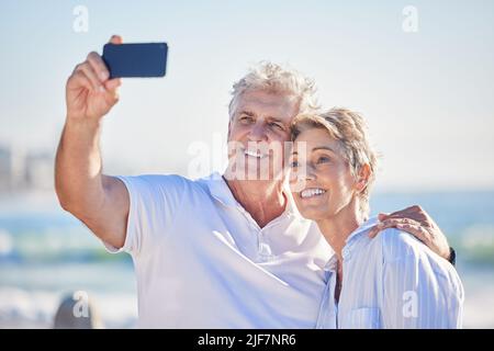 A happy mature caucasian couple enjoying fresh air on vacation at the beach while using a cellphone. Smiling retired couple hugging and taking selfies Stock Photo