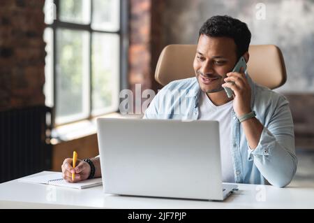 Smiling indian freelancer guy in casual wear sits at the desk in modern office, using trendy laptop for a work, talking on the smartphone and taking notes, making schedule, planning meeting Stock Photo