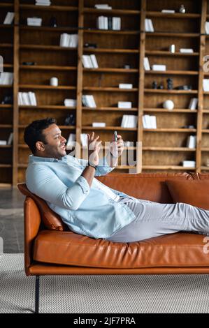 Glad to meet. Vertical side view of cheerful happy indian man in casual wear using smartphone app for virtual communication, holds phone in hand and waving at webcam, holding video call lying on sofa Stock Photo