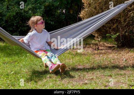 One single school age child, girl wearing cool sunglasses sitting on a hammock, resting in the sun summer leisure activities outdoors lifestyle scene. Stock Photo