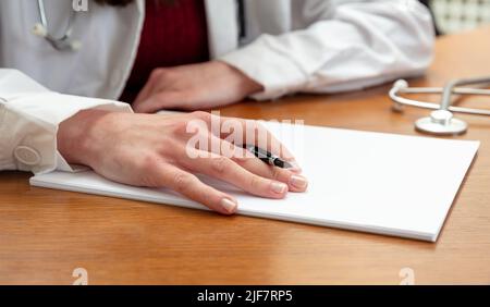 Woman doctor holding a pen on a blank white paper, close up view, wooden office table background. Female hand over empty document Stock Photo