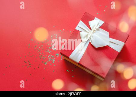 Defocused round lights in the foreground. Beautiful red gift box with satin light ribbon on red background. Green stars shine on the table. Gift for Christmas and New Year. Copy space Stock Photo