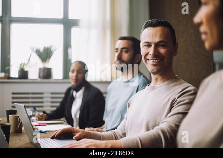 Happy mature businessman with laptop sitting by colleagues in meeting room Stock Photo