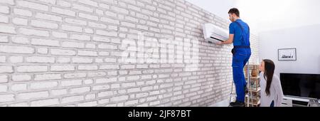 Young Woman Looking At Male Technician Fixing Air Conditioner On Brick Wall Stock Photo