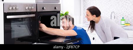 Smiling Young Woman Looking At Technician Fixing Oven In Kitchen Stock Photo