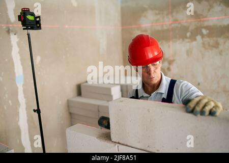 Bricklayer using rubber mallet hammer to tap and level concrete blocks wall. Contractor uses precise spirit bubble and laser level for brickwork Stock Photo
