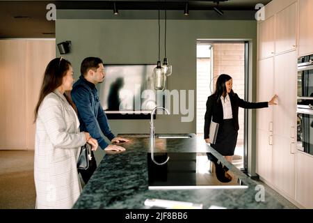Real estate agent showing kitchen cabinets of new house to couple Stock Photo