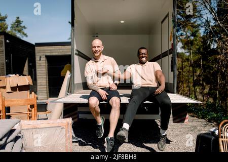 Portrait of smiling multiracial movers sitting in trunk of delivery truck Stock Photo