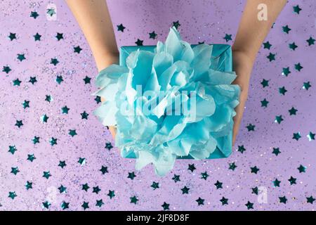A gift wrapped in blue eco kraft paper with a lush bow on a lilac background with small confetti in the form of stars.The holiday, surprise, birthday. Stock Photo