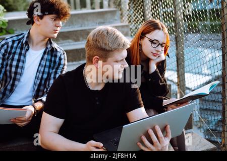 Three friends studying with laptop outdoors sitting on stairs. Small group of high school people preparing for exam.  Stock Photo