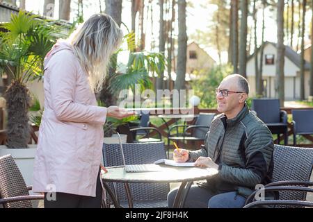 Portrait of smiling middle-aged man father writing down notes in notepad, listening to young woman daughter in cafe. Stock Photo