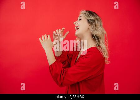 Portrait side view of cheerful young woman in red outfit playing on invisible instrument isolated on red background. Sing and speak into mouthpiece Stock Photo