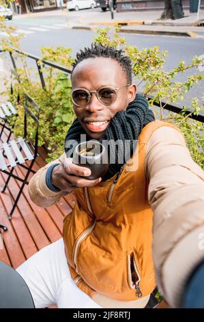 young african man wearing glasses, scarf and winter clothes, makes selfie with his phone drinking coffee sitting outside coffee shop, looking at camer Stock Photo