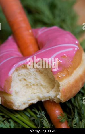 Fresh carrot in pink glazed doughnut with bite taken out, on bed of carrot greens. Stock Photo