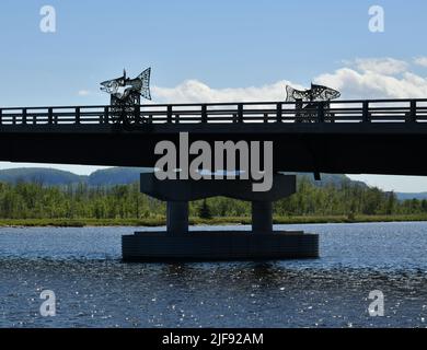 A bridge with iron artwork spans a river in Thunder Bay, Ontario, Canada, while green hills in the background. Stock Photo