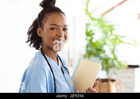 Portrait of smiling african american female doctor with medical report standing against wall at home Stock Photo