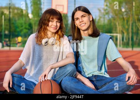 Friends teenagers guy and girl looking at camera, sitting on basketball court Stock Photo