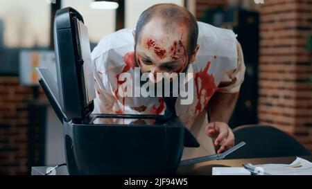 Stupid zombie with blood on clothes photocopying his own face while in office. Creepy doomsday walking dead corpse with bloody wounds damaging photocopy electronic machine in company workspace Stock Photo