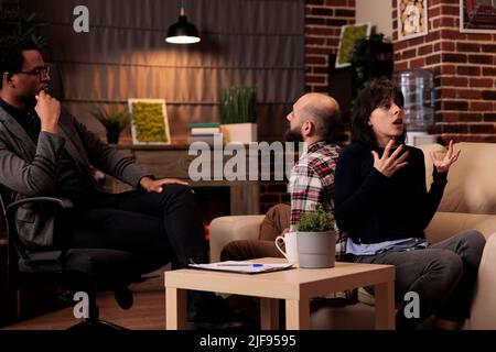 Husband and wife fighting at couple therapy session with counselor, sitting back toback to talk about problems. Therapist giving advice, trying to solve relationship issues between partners. Stock Photo