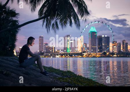 Lonely man sitting under palm tree and using phone against illuminated urban skyline at twilight. Stock Photo